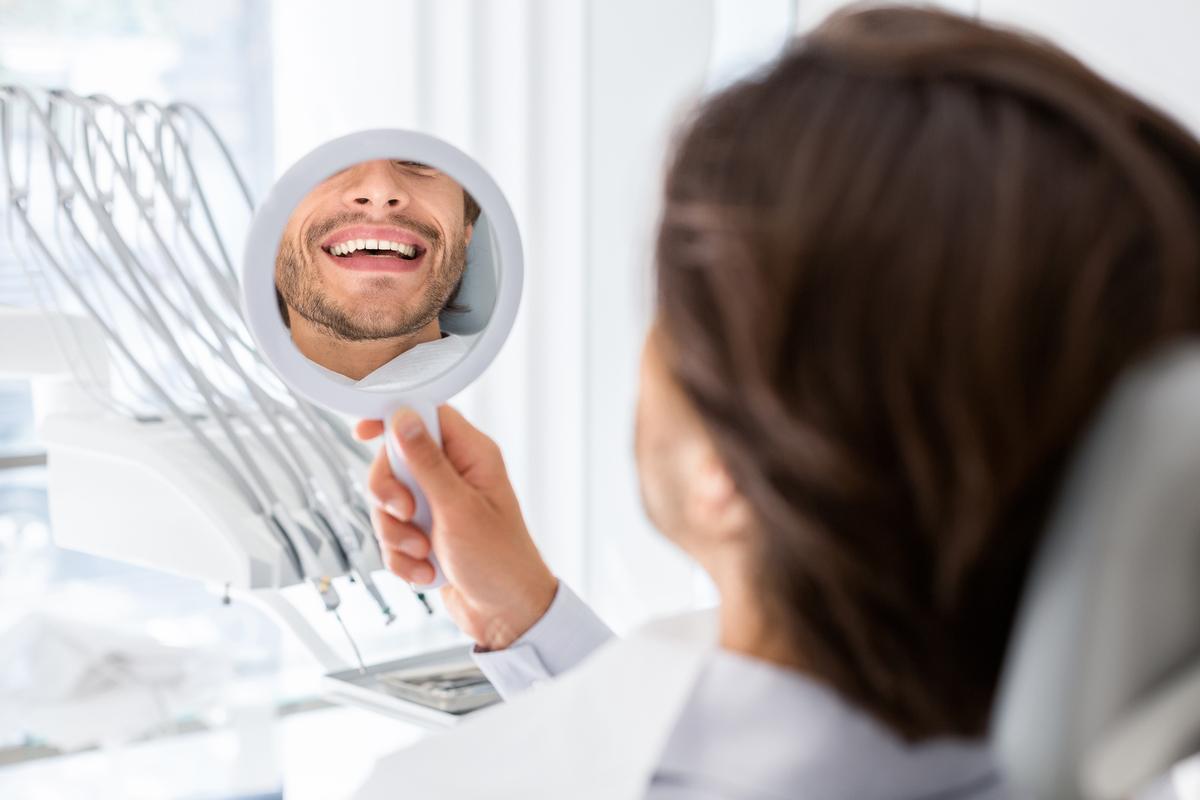 Satisfied man looking at mirror and checking his teeth in dental clinic, beautiful smile concept, cropped